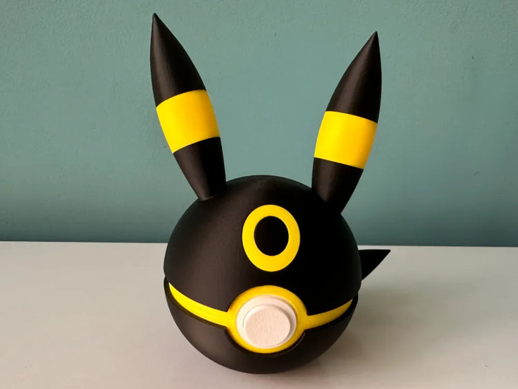 Bola Umbreon negra con detalles amarillos, orejas alargadas y círculo blanco frontal.